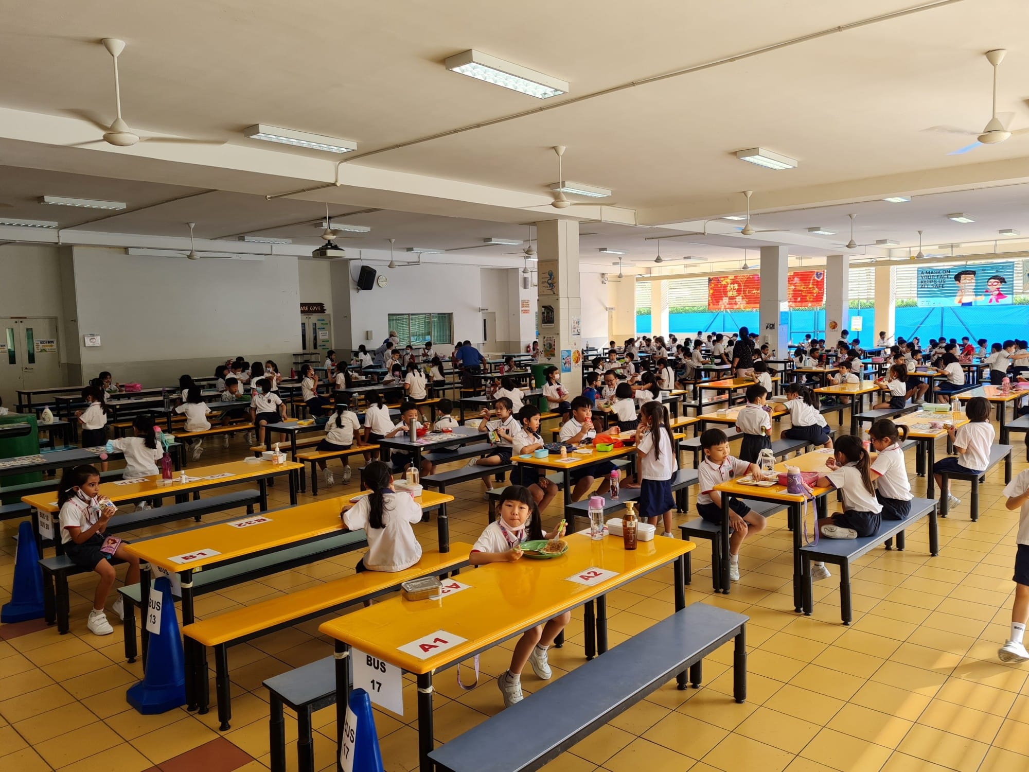 School children in white uniforms eating lunch at yellow tables in a large school canteen with ceiling fans.